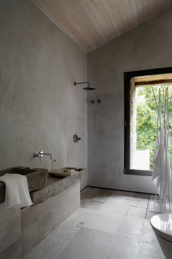 a grey wabi-sabi bathroom with concrete walls, grey tiles, stone sinks and a large window to enjoy the views