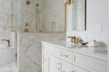 a large white bathroom clad with various types of tiles, with gold handles, fixtures, framing and sconces is a chic space