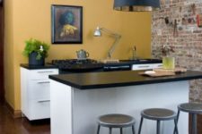 a mini kitchen space with a dark yellow wall, white cabinets, black countertops, a black pendant lamp and a vintage artwork