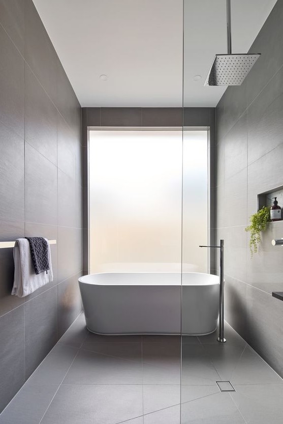 a minimalist bathroom clad with grey large scale tiles, a frosted glass window and some greenery