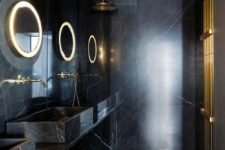 a minimalist black and gold bathroom clad with black marble, with lit up mirrors, gold accessories and fixtures