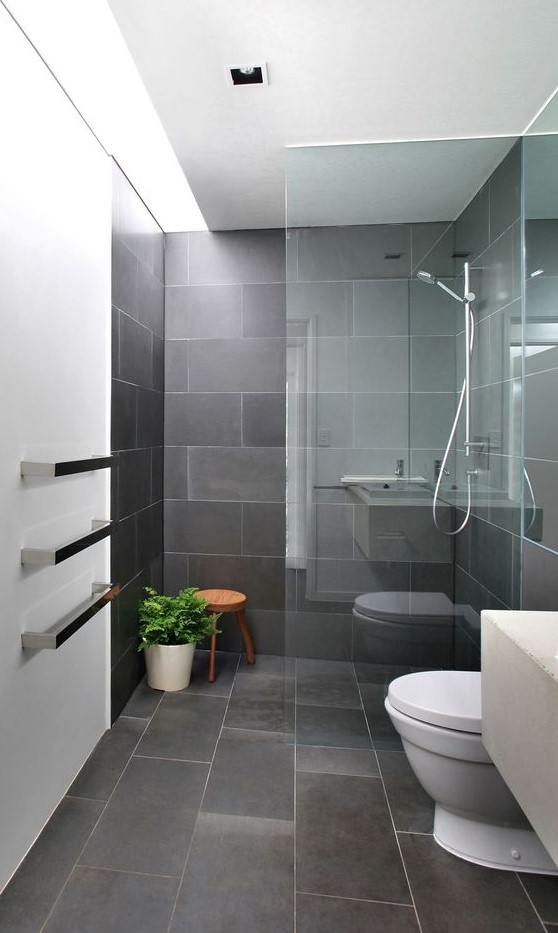 a minimalist grey bathroom clad with tiles, with a glass enclosed shower, white appliances and potted greenery