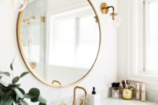 a neutral bathroom with gold fixtures, gold sconces and a round gold framed mirror plus smaller accessories in gold