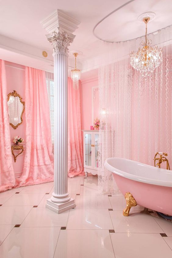 a refined and glam pink bathroom with curtains and lace curtains, a pink bathtub on gold legs, a glam crystal chandelier and a pillar