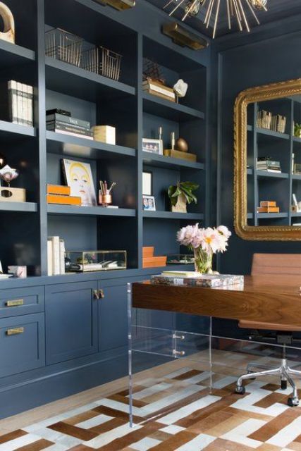 a refined home office with navy walls taken by storage units, with an acrylic and wooden desk, touches of gold for chic and style