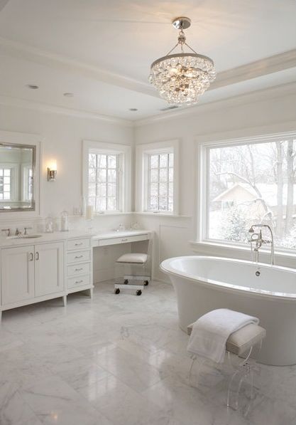 a refined white bathroom with marble tiles, a refined chandelier, a large vanity and makeup nook and windows for light and views