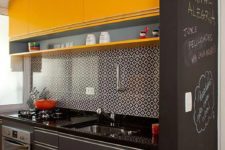 a small black and yellow kitchen with bright upper cabinets, lower grey ones, a mosaic tile backsplash and a chalkboard wall to make notes