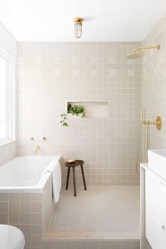 a stylish modern bathroom clad with square grey tiles, a bathtub clad with them, too, a white vanity with a white sink and gold touches