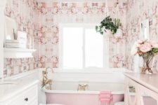 a stylish vintage-inspired bathroom done with pink printed wallpaper, a pink bathtub, a pink sideboard and brass fixtures and legs
