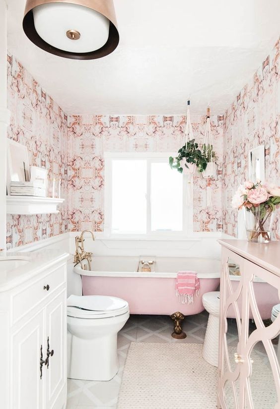 a stylish vintage-inspired bathroom done with pink printed wallpaper, a pink bathtub, a pink sideboard and brass fixtures and legs