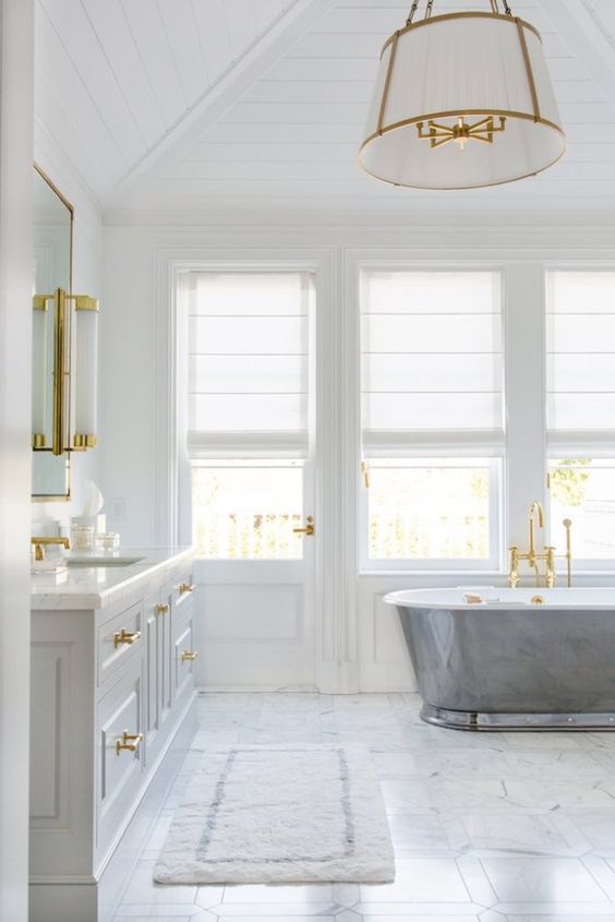 a white bathroom with gold sconces, gold handles, fixtures and a lamp with gold accents is airy and serene