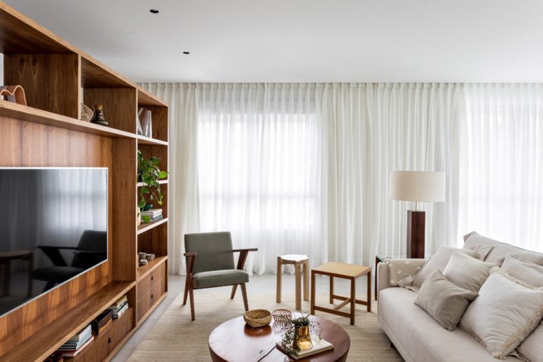 The living room is done with a large wooden unit for storage that divides the spaces, some comfy modern furniture and airy curtains