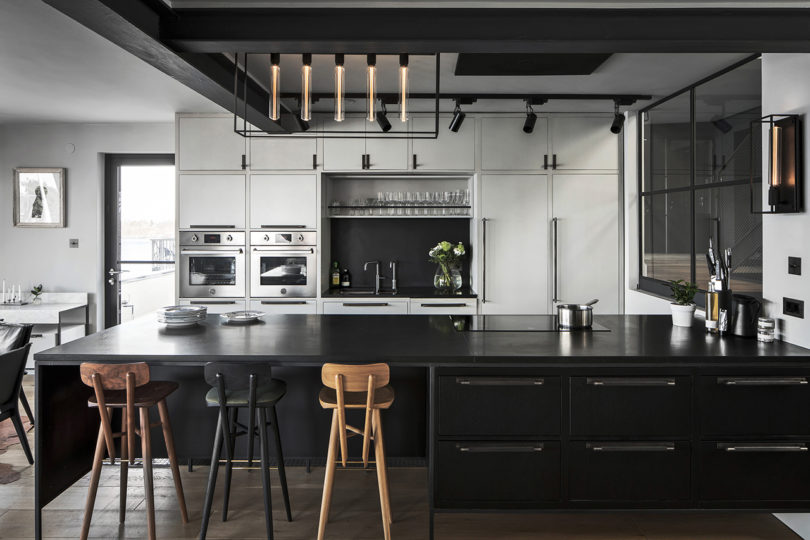 The kitchen is black and white, with plywood and metal cabinets, with a catchy lamp and tall stools