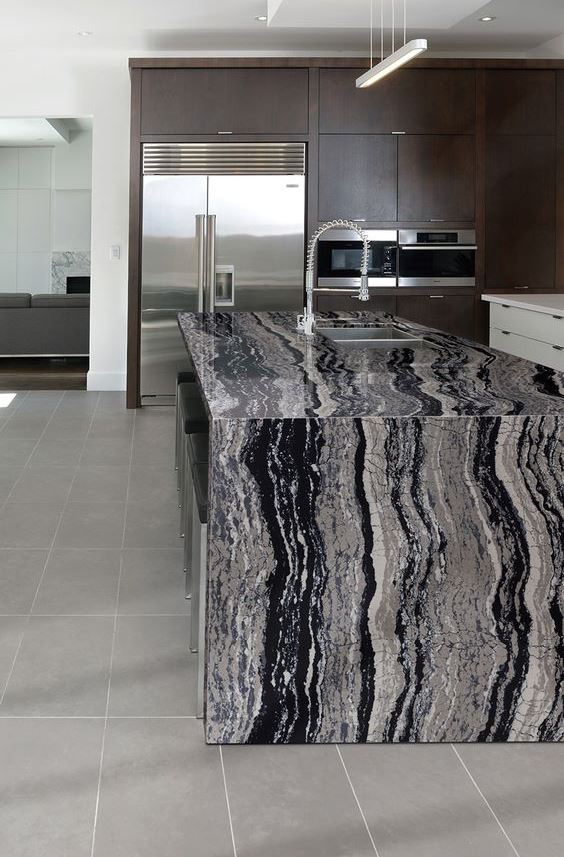 a dark kitchen with a catchy kitchen island and a bold waterfall stone countertop plus stainless steel appliances for a modern feel