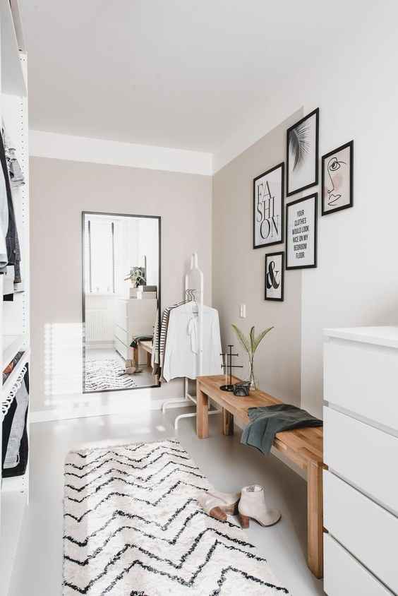 a small yet chic closet in neutrals, with a wooden bench, a mirror, a dresser, holders with hangers and a gallery wall