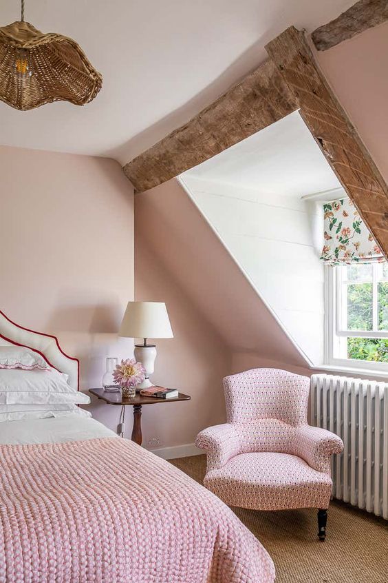 a blush attic bedroom with a white upholstered bed and pink and white bedding, a pink chair, a nightstand with a table lamp
