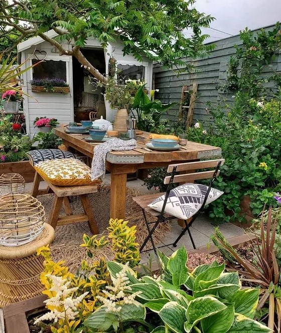 a bright and cool backyard with a lot of greenery, a wooden dining set and folding chairs, candle lanterns and blooms