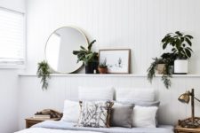 a chic coastal bedroom with a white beadboard wall, wooden furniture, potted plants and greenery, a round mirror and comfy textiles