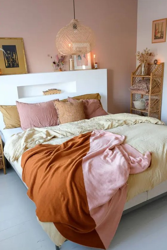 a lovely bedroom with a pink accent wall, a bed with an accent headboard, a pendant lamp and pretty bedding, a woven shelf and some artworks