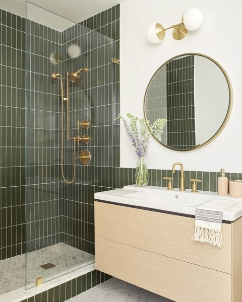 a modern bathroom with green skinny tiles, a floating vanity, a round mirror, gold and brass fixtures is cool