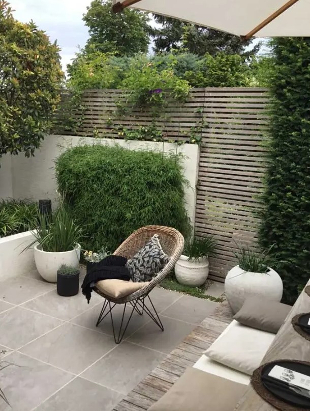 a modern paved patio with a planked screen, with a tiled floor, potted grasses and greenery, a small dining set with pillows and a woven chair