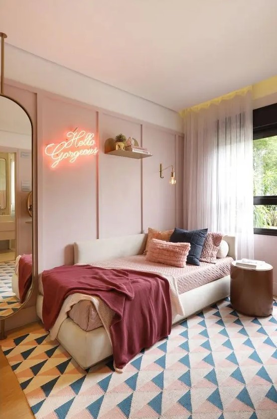 a pale pink paneled accent wall, a creamy daybed with comfy pillows, a neon sign, a large mirror and a round side table