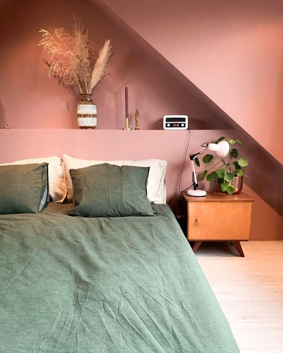 a pink attic bedroom with a built-in shelf with decor, a bed with green bedding, a stained nightstand and greenery