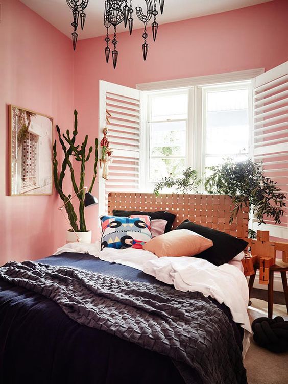 a pink bedroom with a bed and a woven headboard, black and white bedding, potted plants, a black chandelier with pendants