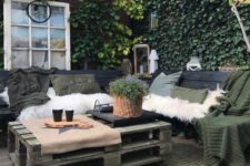 a small Nordic patio with pallet furniture, dark textiles, faux fur covers, pendant lamps, potted greenery and a fire pit