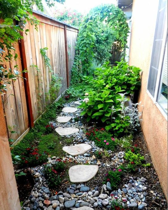 a small and bright garden nook with pebbles, greeneyr and bright blooms, shrubs and a climbing plants on the fence