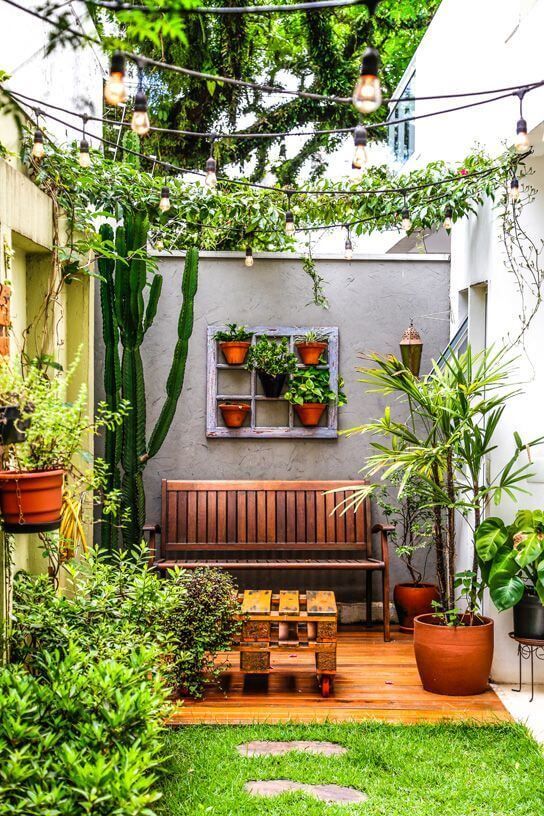 a small and cool patio with greeneyr, potted cacti and plants, a wooden bench and a pallet table, some lamps around