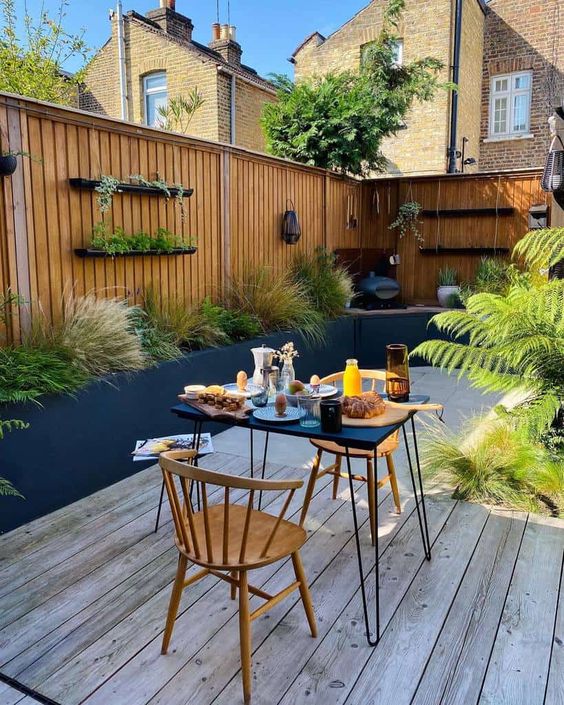 a small and cool pation with a wooden deck, greenery and grasses, a hairpin table and wooden chairs, some shelves is awesome