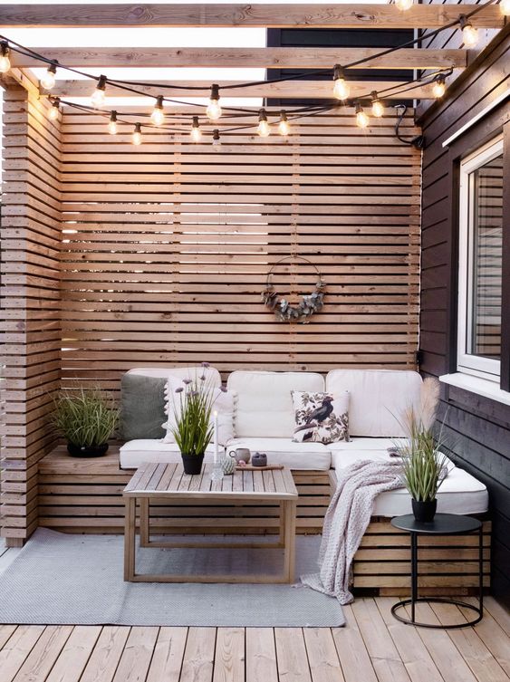a small and cute patio with wood slat privacy screens and a bench, neutral cushions, a coffee table, string lights and potted greenery