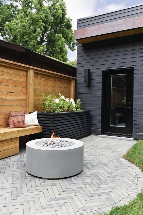 a small and laconic patio with a wooden bench, a tall black planter with greenery and blooms and a fire pit is a cool space