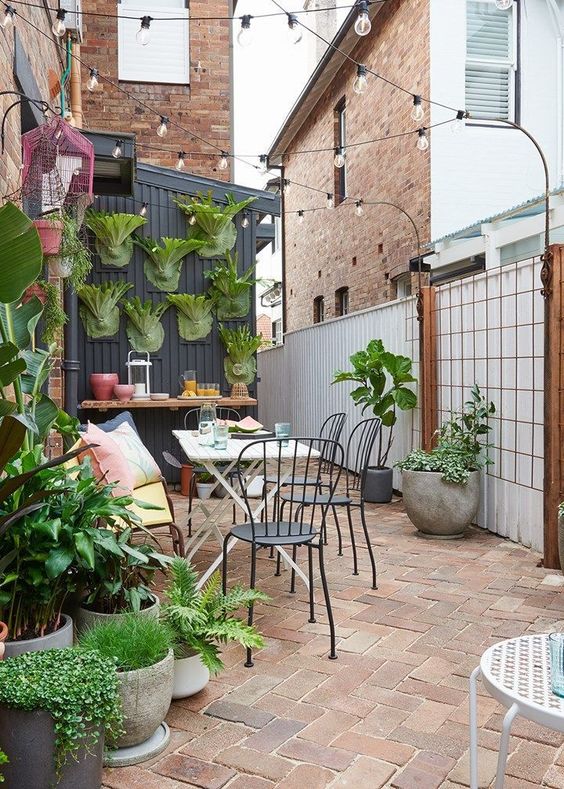 a small and lovely patio with potted greenery, a bar counter, a wooden table and black chairs and some string lights over the space