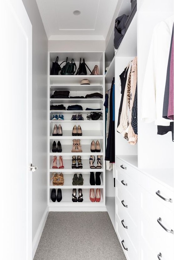 a small and narrow white walk-in closet with holders for clothes, open shelves for shoes, drawers for smaller stuff is cool and organized