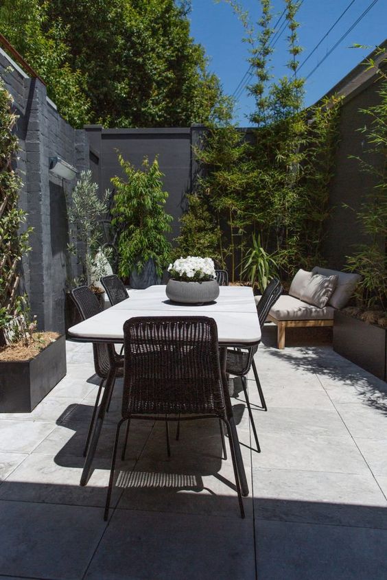 a small and stylish modern patio with a large table and black chairs, a daybed, greenery surrounding the space