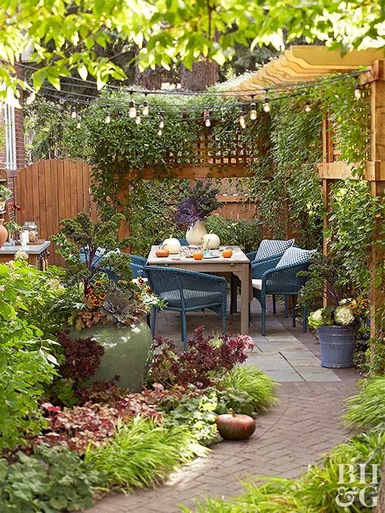 a small and welcoming patio with greenery, a wooden table and woven chairs, lots of greenery and blooms, lovely and cozy fall decor