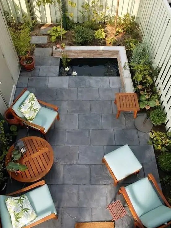 a small and zen-like paved patio with tiles on the ground, a tiny pond, some growing plants and cool garden furniture with blue upholstery