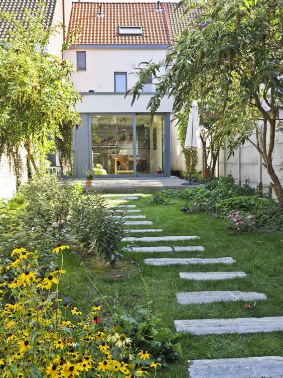 a small backyard gardem with a green lawn, greenery and bright blooms and some trees plus a pavement