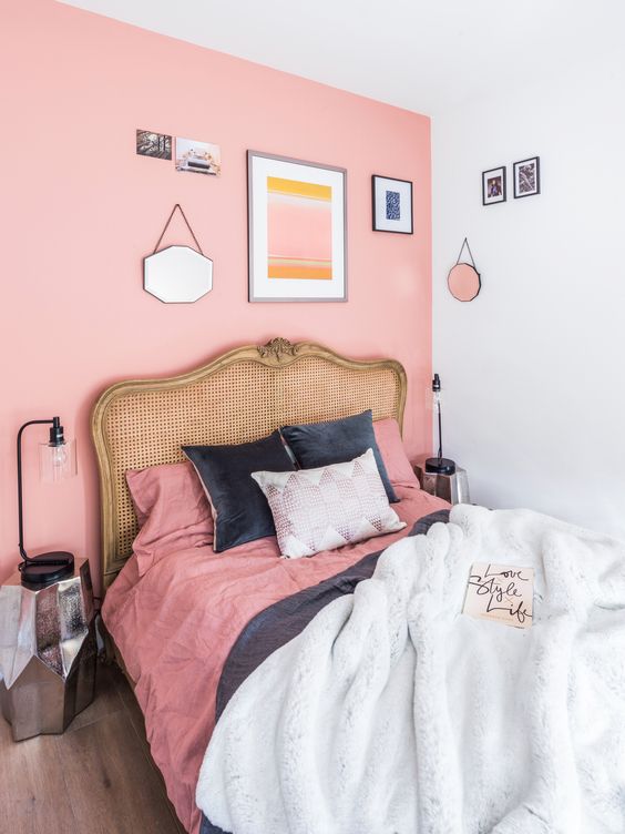 a small bedroom with a pink accent wall, a bed with a cane headboard and pink and black bedding, a gallery wall and mirror faceted nightstands