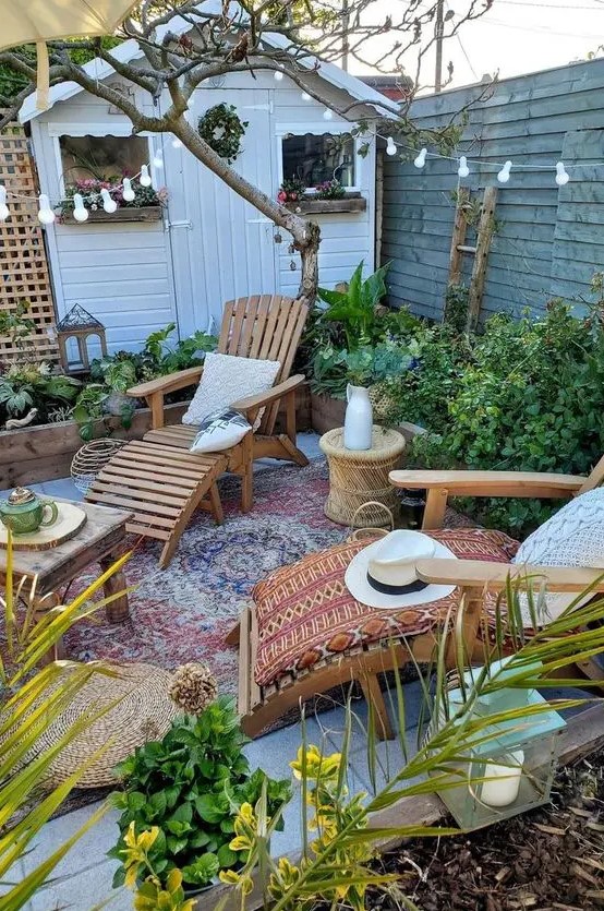 a small boho backyard with a boho rug, wooden loungers, some tables, greenery and blooms around, string lights and jute poufs