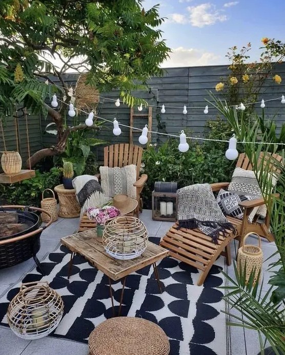 a small boho backyard with a wodoen deck, wooden loungers, a table, some jute poufs, candle lanterns, string lights and greenery