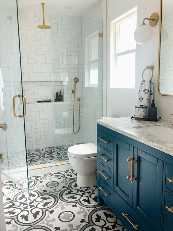 a small bright bathroom with white tiles in the shower, printed tiles on the floor, a navy vanity and brass fixtures