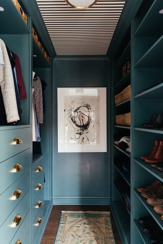 a small closet done in blue, with elegant gold knobs, open storage space, drawers and a gold lamp on the striped ceiling