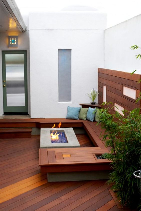 a small contemporary patio clad with wood, with a built-in wooden bench, a fire pit, some potted greenery and pillows