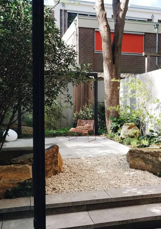a small contemporary patio with a stone deck, some trees, greenery and plants, large rocks and pebble lamps on the ground