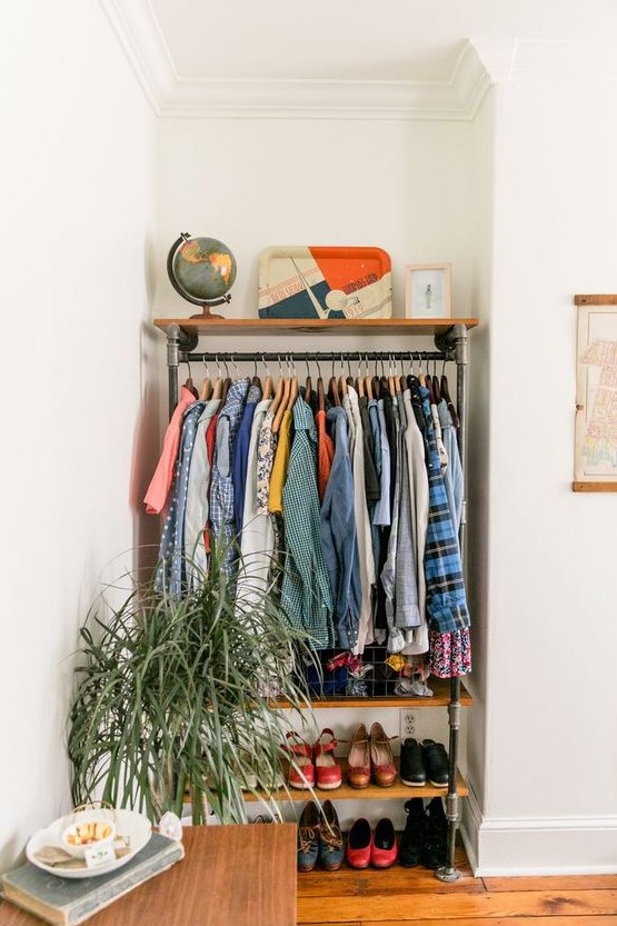 a small corner makeshift closet with a metal and wood shelf, a potted plant and lots of colorful clothes plus a dresser