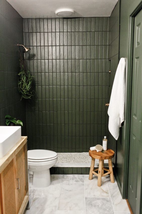 a small green bathroom with skinny tiles, a cane vanity, a wooden stool and some plants and white towels