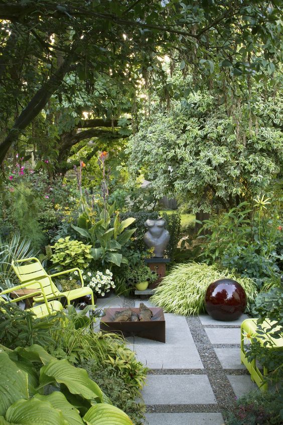 a small lush garden with lots of grasses, shrubs and flowers, some trees, a fire pit and garden chairs
